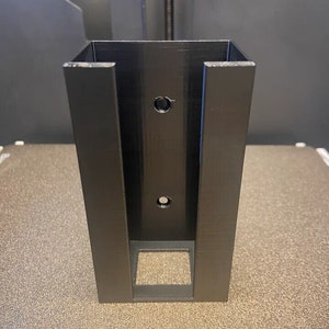 Op de afbeelding: Een zwarte plastic wandbeugel met twee gaten en een rechthoekige opening. De beugel is ontworpen om een klein object te houden, zoals een telefoon of een luidspreker.