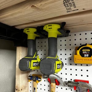 May include: Two black and neon yellow Ryobi cordless drills suspended from a wooden shelf. Below, a pegboard displays various tools, including hammers and a yellow tape measure. The tools are organised for easy access in a workshop.