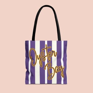 Puede incluir: Bolso tote con un diseño de rayas moradas y blancas. Las palabras "Oyster Bay" están escritas en letras doradas. El bolso tiene asas negras y probablemente está hecho de tela.