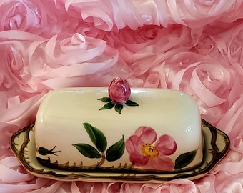 May include: A white ceramic butter dish with a floral design. The dish has a lid with a pink rosebud on top. The dish is decorated with green leaves and pink roses.