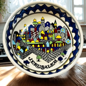 May include: A round ceramic bowl featuring a hand-painted, colorful cityscape of Jerusalem. The design includes intricate black line work, vibrant yellow, red, and blue buildings, and a thick, wavy dark blue border. The word JERUSALEM is printed.