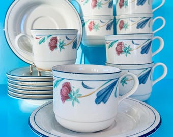 May include: A set of white teacups and saucers with a floral design. The cups and saucers have a blue trim and a pattern of red and blue flowers. The set includes multiple cups, saucers, and a serving plate, all arranged on a blue surface.