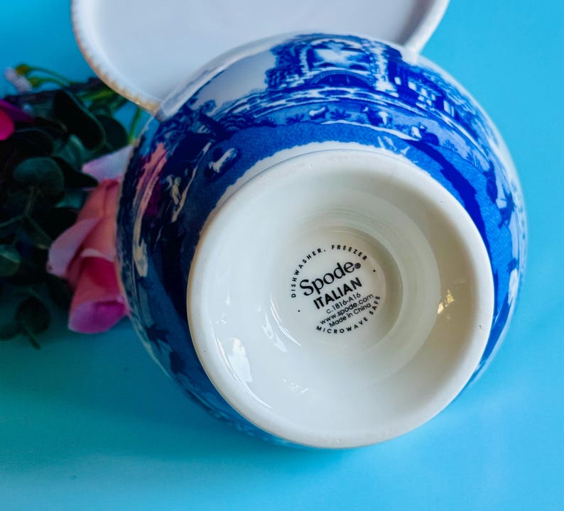 May include: A white and blue Spode Italian teacup with a handle. The teacup features a detailed blue landscape design against a white background. The bottom of the teacup has the Spode Italian logo and product information.