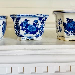 May include: Three white ceramic planters with blue floral designs. The planters are different sizes and shapes, including square and octagonal. The blue floral patterns are detailed and cover the sides of the planters. The planters are sitting on a white shelf.