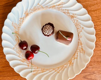 May include: A white, scalloped-edge plate with gold filigree holds three dark red cherries and two chocolates. One chocolate is round with a lattice design, the other is rectangular with a pink flower.