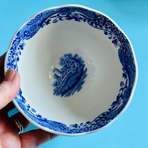May include: A white ceramic bowl with a blue floral and landscape design. The rim has a wavy edge and is decorated with a blue floral pattern. The interior features a blue landscape scene. The bowl is held against a light blue background.