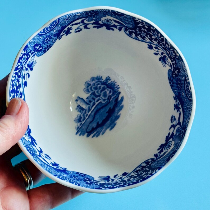 May include: A white ceramic bowl with a blue floral and landscape design. The rim has a wavy edge and is decorated with a blue floral pattern. The interior features a blue landscape scene. The bowl is held against a light blue background.