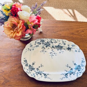 May include: An oval, white ceramic serving platter with a scalloped edge. The platter is decorated with a blue floral pattern. A small bouquet of colorful flowers sits in a silver vase in the background.