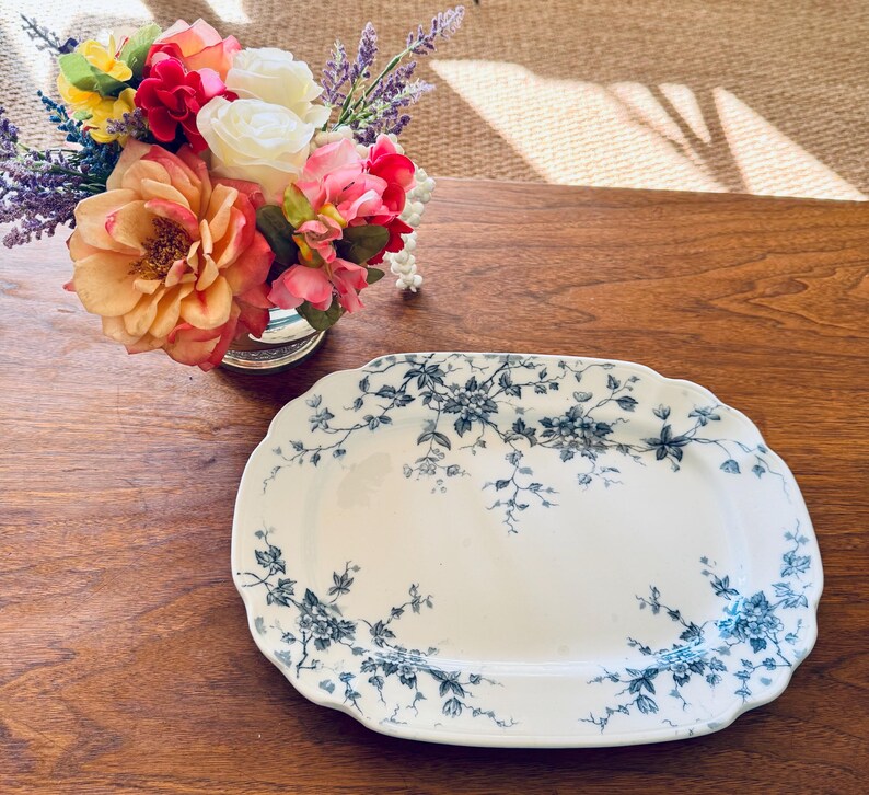 May include: An oval, white ceramic serving platter with a scalloped edge. The platter is decorated with a blue floral pattern. A small bouquet of colorful flowers sits in a silver vase in the background.