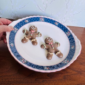 May include: Two chocolate gingerbread men on a white plate with a blue floral border. The gingerbread men have icing details, including smiles, bow ties, and green and tan accents. The plate is held by a hand.