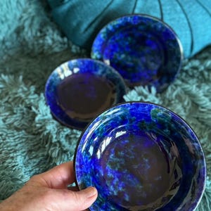 May include: Three round ceramic bowls featuring a glossy finish with a mottled pattern of deep cobalt blue, navy, and dark green hues. The bowls have a smooth, reflective surface and a circular shape, with one bowl held in the foreground.