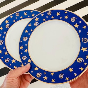 May include: Two white ceramic plates with a blue border featuring gold stars and swirls. The plates have a gold trim and are held against a black and white striped background. The plates are for serving food.