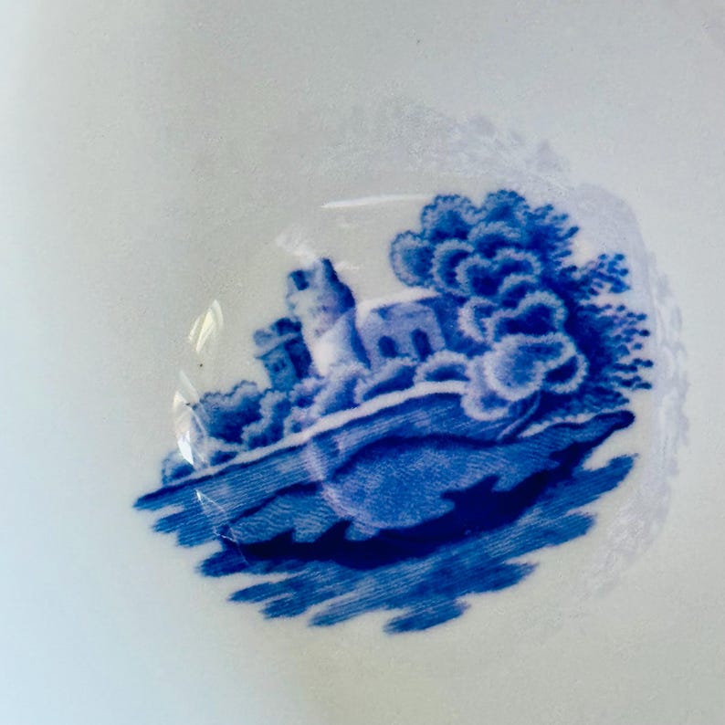 May include: A close-up of a white ceramic surface with a blue and white transferware design. The design depicts a castle scene with trees and a body of water. The image is a close-up of a portion of the design.