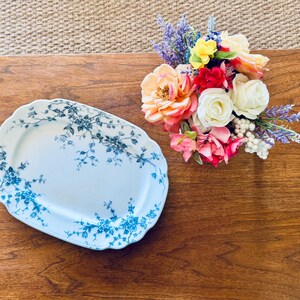 May include: An oval, white serving platter with a blue floral design. A bouquet of colorful flowers, including roses and lavender, sits to the right of the platter. The items are on a wooden surface.