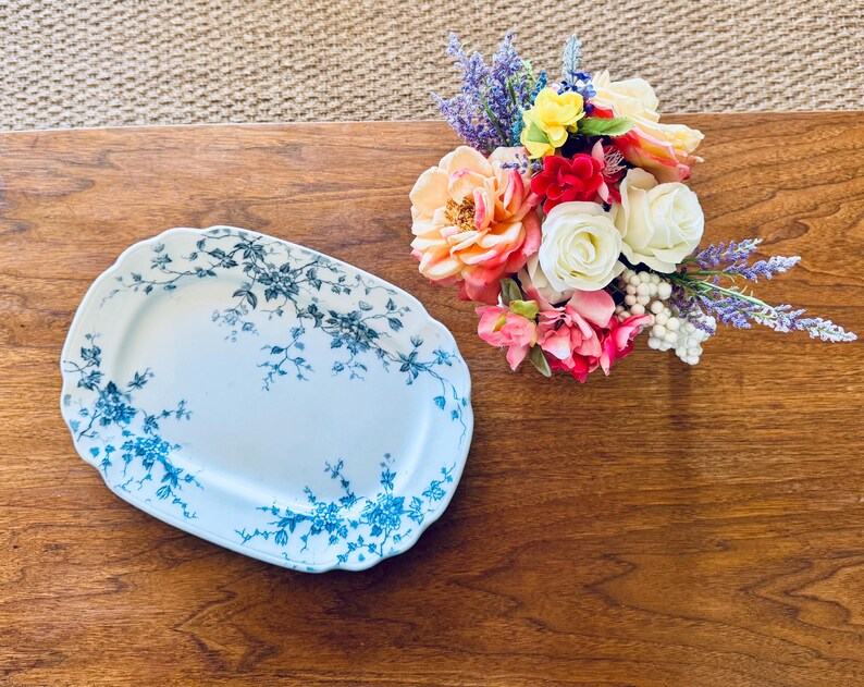 May include: An oval, white serving platter with a blue floral design. A bouquet of colorful flowers, including roses and lavender, sits to the right of the platter. The items are on a wooden surface.