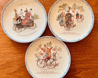 May include: Three vintage decorative plates with illustrations of people and vehicles. Each plate features a different scene with people in antique cars and bicycles. The plates have a white background with a blue patterned border and text.