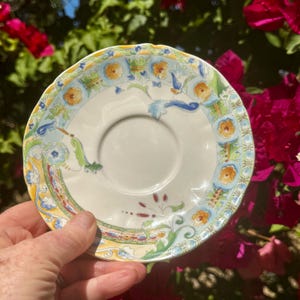 May include: A white ceramic saucer with a scalloped edge. The saucer features a colorful floral design with orange, blue, and green accents. The design includes stylized flowers and foliage, creating a vintage aesthetic.