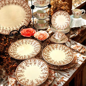 May include: A collection of cream-colored ceramic dinnerware featuring a brown floral pattern. The set includes a large platter, several dinner plates, a three-tier stand with cups and saucers, a creamer pitcher, and individual teacups with saucers.