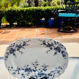 May include: A white, oval serving platter with a scalloped edge, adorned with a blue floral pattern. The platter is displayed on a black metal stand, showcasing its elegant design. The background is a sunny outdoor setting.