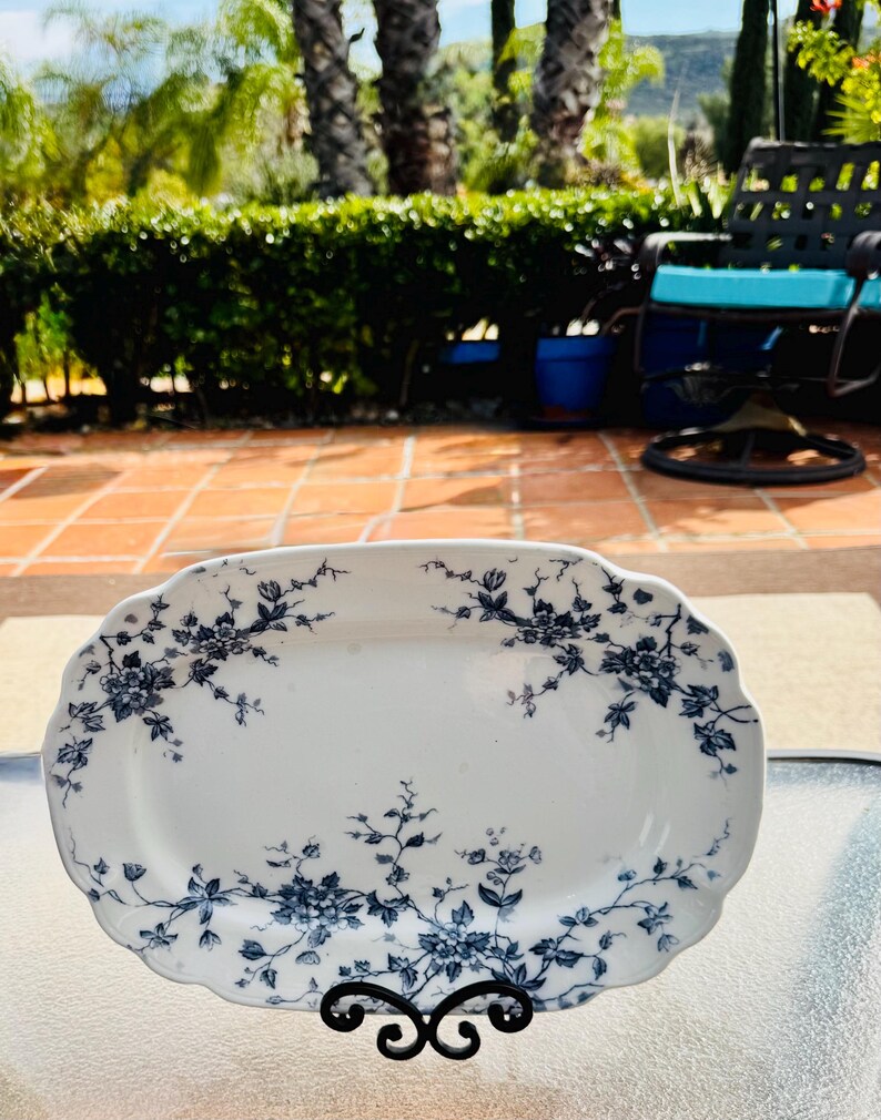 May include: A white, oval serving platter with a scalloped edge, adorned with a blue floral pattern. The platter is displayed on a black metal stand, showcasing its elegant design. The background is a sunny outdoor setting.