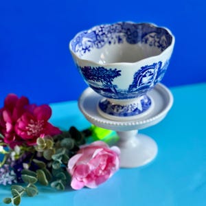 May include: A white and blue ceramic footed bowl with a detailed landscape design. The bowl sits on a white pedestal, with a blue background. Pink and purple flowers are in the foreground.