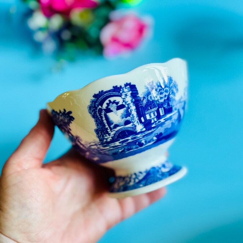 May include: A white and blue ceramic bowl with a pedestal base. The bowl features a blue transferware design of a landscape scene. The rim has a scalloped edge. The bowl is held in a hand against a blue background.