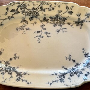 May include: An antique, rectangular serving platter with a cream-colored base. The platter is decorated with a floral pattern in shades of gray and blue. The flowers and vines are arranged around the edges of the platter.