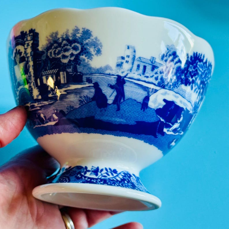 May include: A white and blue ceramic footed bowl with a scalloped rim. The bowl features a blue landscape design with figures and buildings. The base of the bowl has a decorative blue floral pattern.