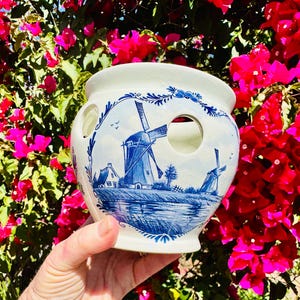 May include: A white ceramic planter with a blue and white windmill design. The planter has two large circular openings and is held in front of a background of bright pink flowers.