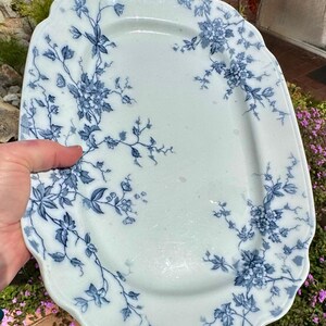 May include: An antique, rectangular serving platter with a scalloped edge. The white ceramic platter features a blue floral pattern with leaves and blossoms. The design is concentrated around the edges and corners of the platter.