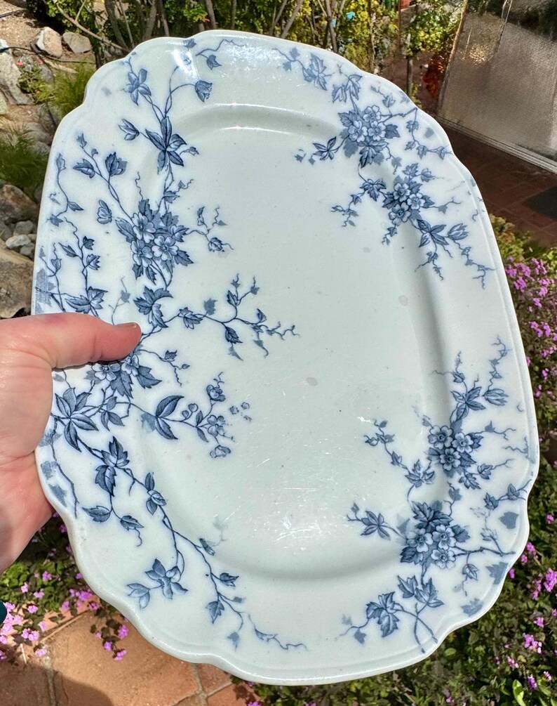 May include: An antique, rectangular serving platter with a scalloped edge. The white ceramic platter features a blue floral pattern with leaves and blossoms. The design is concentrated around the edges and corners of the platter.