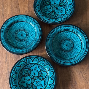 May include: Four round ceramic plates featuring a vibrant turquoise and black color scheme. Two plates display a central geometric lattice pattern, while the other two feature a symmetrical floral-inspired motif with intricate black line work.
