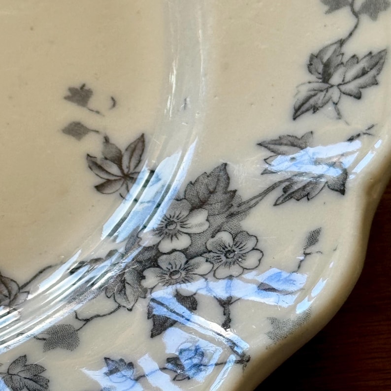 May include: Cream-colored ceramic plate with a floral design. The plate features a scalloped edge and is decorated with gray and white flowers and leaves. The design is repeated around the rim of the plate.