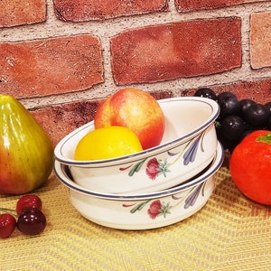 May include: Two white ceramic bowls with blue and green floral designs. The bowls are stacked on top of each other. The top bowl contains a peach and a yellow lemon. The bottom bowl is empty.