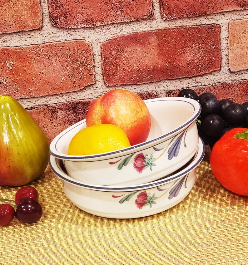 May include: Two white ceramic bowls with blue and green floral designs. The bowls are stacked on top of each other. The top bowl contains a peach and a yellow lemon. The bottom bowl is empty.