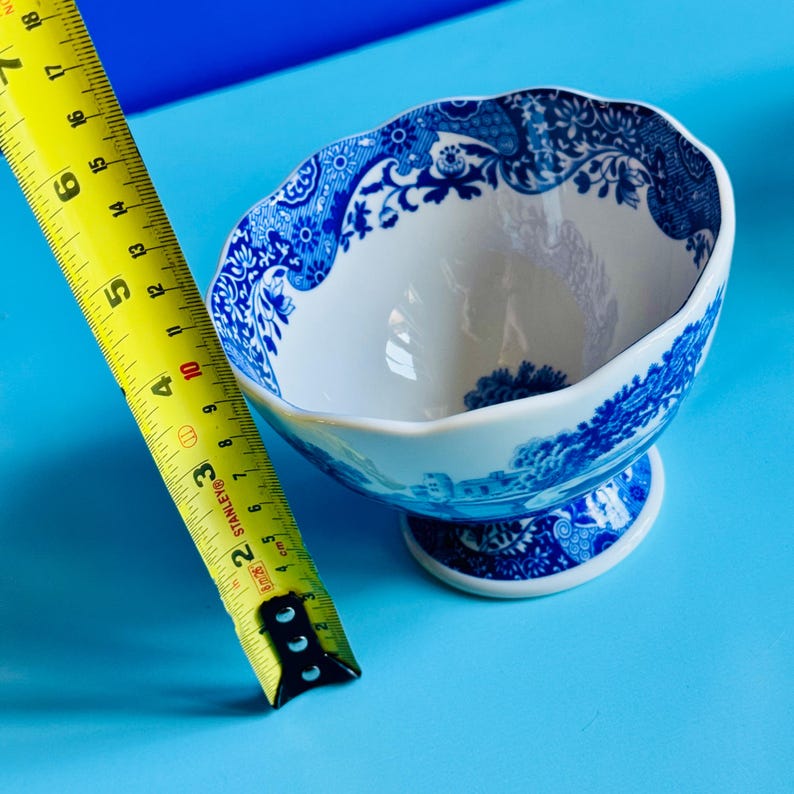 May include: A white and blue ceramic footed bowl with a decorative floral and landscape pattern. The bowl is approximately 4 inches in diameter. The bowl is on a blue surface.
