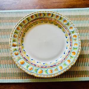 May include: A white ceramic plate with a decorative border featuring a repeating floral pattern in yellow, blue, and green. The plate is set on a green and brown striped placemat, creating a colorful and inviting presentation. The plate's design adds a touch of elegance.