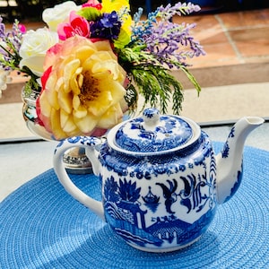 May include: A blue and white ceramic teapot with a Willow pattern design. The teapot has a curved handle and spout, and a matching lid. A bouquet of colorful flowers sits in a silver vase in the background.