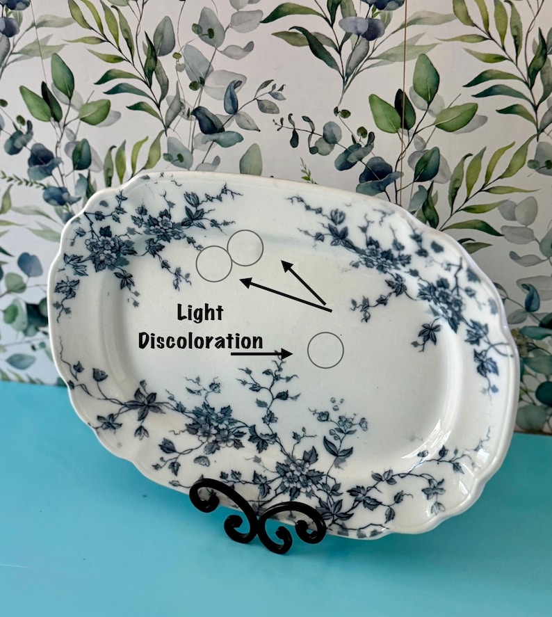 May include: An oval, white ceramic serving platter with a blue floral design. The platter has a scalloped edge and is supported by a black metal stand. Text on the platter indicates light discoloration.