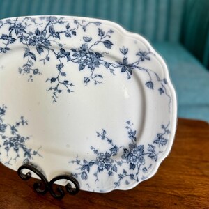 May include: White oval serving platter with a scalloped edge, adorned with a blue floral pattern. The platter is displayed on a black metal stand, showcasing its elegant design. A classic piece for serving food.