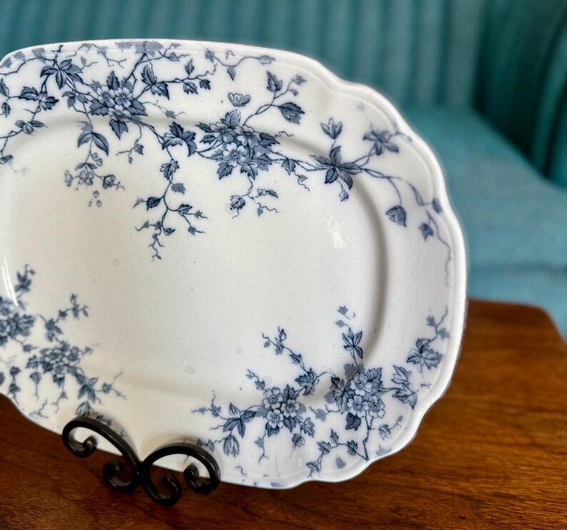 May include: White oval serving platter with a scalloped edge, adorned with a blue floral pattern. The platter is displayed on a black metal stand, showcasing its elegant design. A classic piece for serving food.
