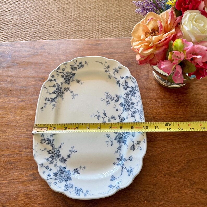 May include: A white, scalloped-edge serving platter with a blue floral design. The platter is approximately 10 inches long. A measuring tape is placed across the platter. A vase of flowers is in the background.