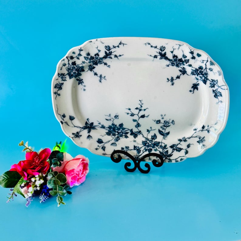 May include: White oval serving platter with a blue floral design. The platter is supported by a black metal stand. A bouquet of pink and red flowers is in the foreground.