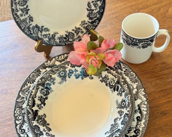 May include: A set of white ceramic dinnerware with a black floral pattern. The set includes a plate, a bowl, and a mug. A pink flower arrangement sits near the dishes. The dishes are on a wooden surface.