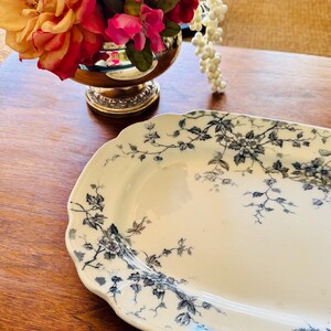 May include: An oval, off-white serving platter with a delicate floral pattern in shades of gray and blue. The edges of the platter are scalloped. The platter is on a wooden surface.