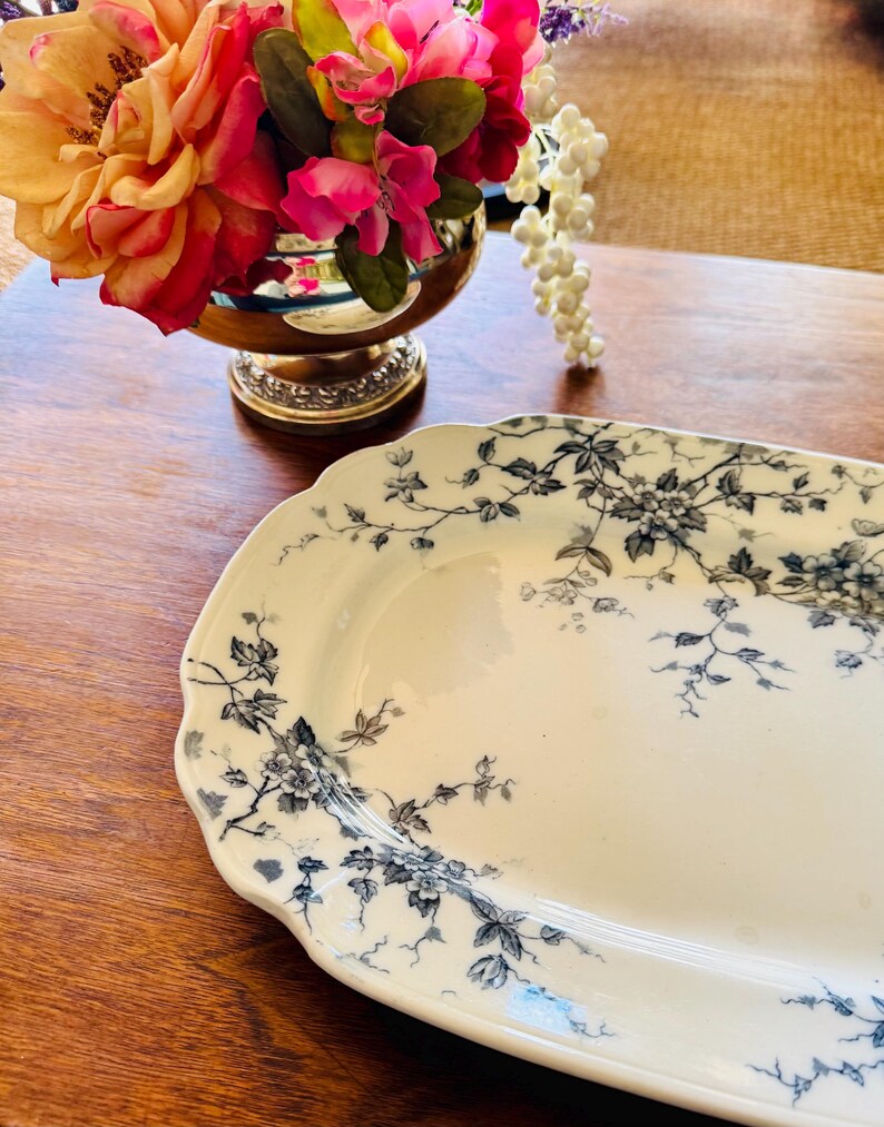 May include: An oval, off-white serving platter with a delicate floral pattern in shades of gray and blue. The edges of the platter are scalloped. The platter is on a wooden surface.