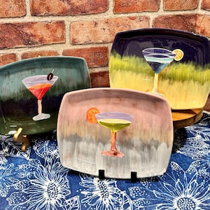 May include: Three square ceramic plates with cocktail designs. Each plate features a different cocktail illustration with a colorful background. The plates are displayed on a blue floral patterned cloth with a brick background.