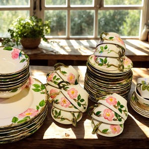 May include: A collection of cream-colored ceramic dinnerware featuring a hand-painted pink rose and green leaf pattern. The set includes stacked dinner plates, bowls, and teacups with matching saucers, all finished with a thin gold-tone rim.