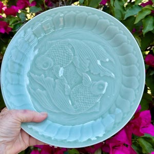 May include: A light blue ceramic plate with a detailed relief of two goldfish swimming among aquatic plants. The plate has a scalloped edge and is held by a hand. The plate's diameter is approximately 10 inches.