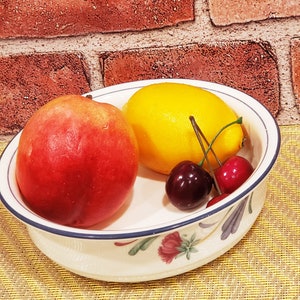 May include: A white ceramic bowl with a blue rim and floral design, containing a red peach, a yellow lemon, and two red cherries.